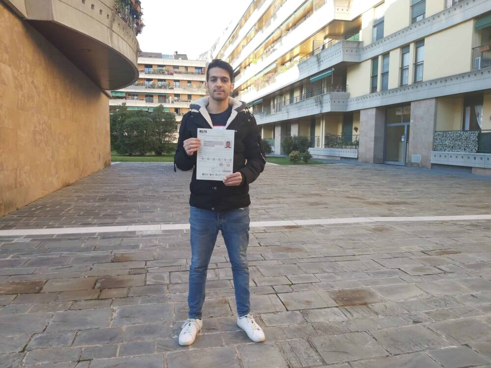 Peyman is holding his IELTS exam report in front of buildings